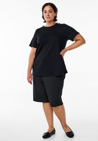 Black cotton t-shirt with a rounded neckline and decorative button accents on one shoulder, paired with black shorts and black flat shoes.