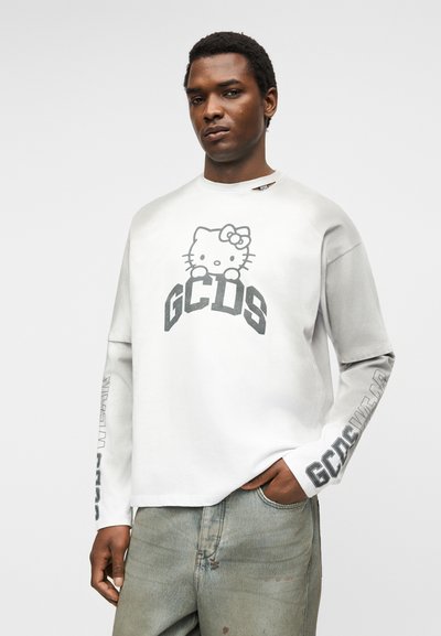 Νέος άνδρας που φοράει λευκό μακρυμάνικο πουκάμισο με γραφικό Hello Kitty και κείμενο "GCDS", σε συνδυασμό με ανοιχτό γκρι τζιν, στέκεται με το χέρι στην τσέπη.