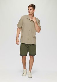 Beige Kurzarmhemd mit strukturierter Musterung, kombiniert mit hochgekrempelten olivgrünen Shorts und cremefarbenen Sneakers. Schlichte Knopfverschlussdetails.