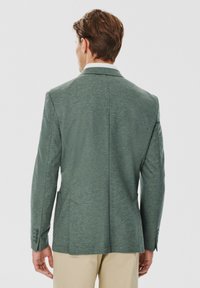 Tył zielonej marynarki z teksturowanej tkaniny, z dwoma tylnymi rozcięciami i mankietami na trzy guziki, w połączeniu z jasnymi spodniami.