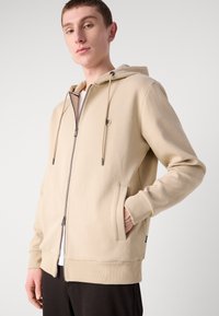 Felpa beige con zip in tessuto morbido, dotata di cappuccio con coulisse, tasche frontali, polsini a coste e una toppa con logo sul petto.