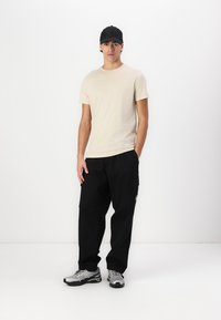 Beidzis īsām piedurknēm krekliņš, melnas cargo bikses, pelēkie sporta apavi. Vīrietis valkā melnu cepuri un stāv uz balta fona.