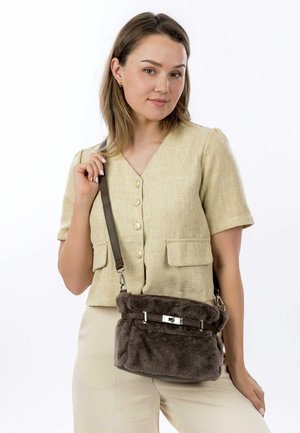 Sac rond en fausse fourrure marron avec une surface texturée, une bandoulière ajustable et un accent de fermoir en argent. Porté par-dessus une chemise verte olive à boutons.