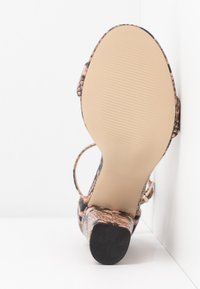 Sandale à talons hauts avec un motif en serpent dans des nuances de beige et de noir. Semelle lisse de couleur crème avec texture nervurée. Design à lanières.