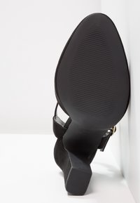 Černá semišová lodička na vysokém podpatku s texturovanou gumovou podrážkou, obsahující blokový podpatek a kotníkový pásek se malou kovovou přezkou.