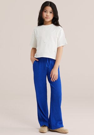 Jauna mergina su ilgais tamsiais plaukais, dėvinti baltą trumpomis rankovėmis megztinį su gėlių detalėmis, ryškias mėlynas tekstūruotas kelnes ir smėlio spalvos batus.