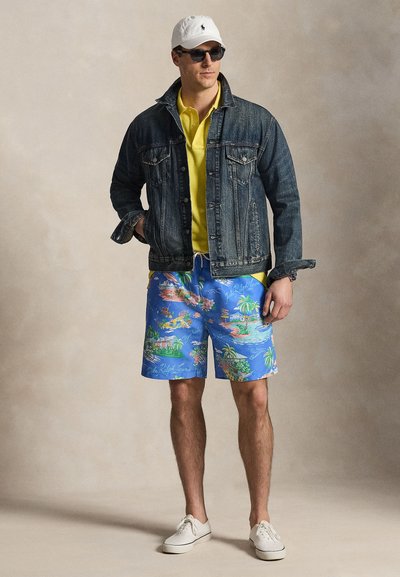 Homme portant une casquette blanche, des lunettes de soleil, un polo jaune, une veste en jean foncé, un short bleu à imprimé tropical et des baskets blanches, debout les mains dans les poches.