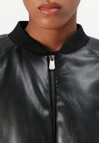 Giacca bomber nera in pelle con colletto a coste, caratterizzata da una zip frontale e una texture liscia. La vista ravvicinata si concentra sulla parte superiore.