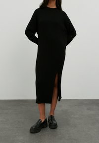 Robe en tricot côtelé noire avec des manches longues et une fente sur le côté, portée avec des mocassins épais noirs, le tout sur fond neutre.