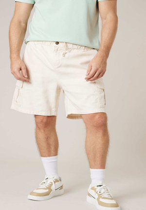 Homme portant un short cargo beige, des chaussettes blanches et des baskets blanches avec des accents beiges, debout devant un fond neutre.
