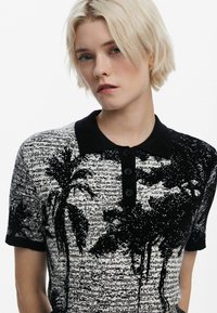 Zwart-wit polo-shirt met een palmbomenpatroon, gebreide structuur en een zwarte kraag met vier knopen.
