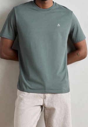 Homme portant un t-shirt vert foncé à col rond avec un petit logo blanc "MOP" sur la poitrine et un pantalon beige clair, debout les mains derrière le dos.