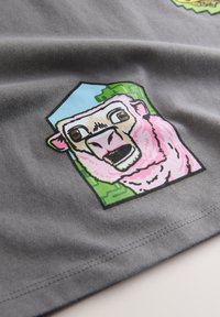 Pelēka auduma t-krekls ar apdruku – pērtiķis ar rozā seju, ko ieskauj zaļas akcentu detaļas uz zilas pamatnes.