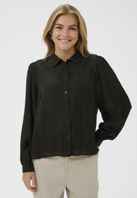 Lächelnde Frau mit blonden Haaren, die eine schwarze Langarmbluse mit Knöpfen und beige Hosen trägt, steht mit einer Hand auf der Hüfte.