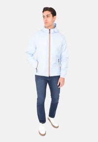 Giacca impermeabile blu chiaro con cappuccio, dotata di una zip arancione e tasche laterali, abbinata a jeans blu scuro e sneakers bianche.