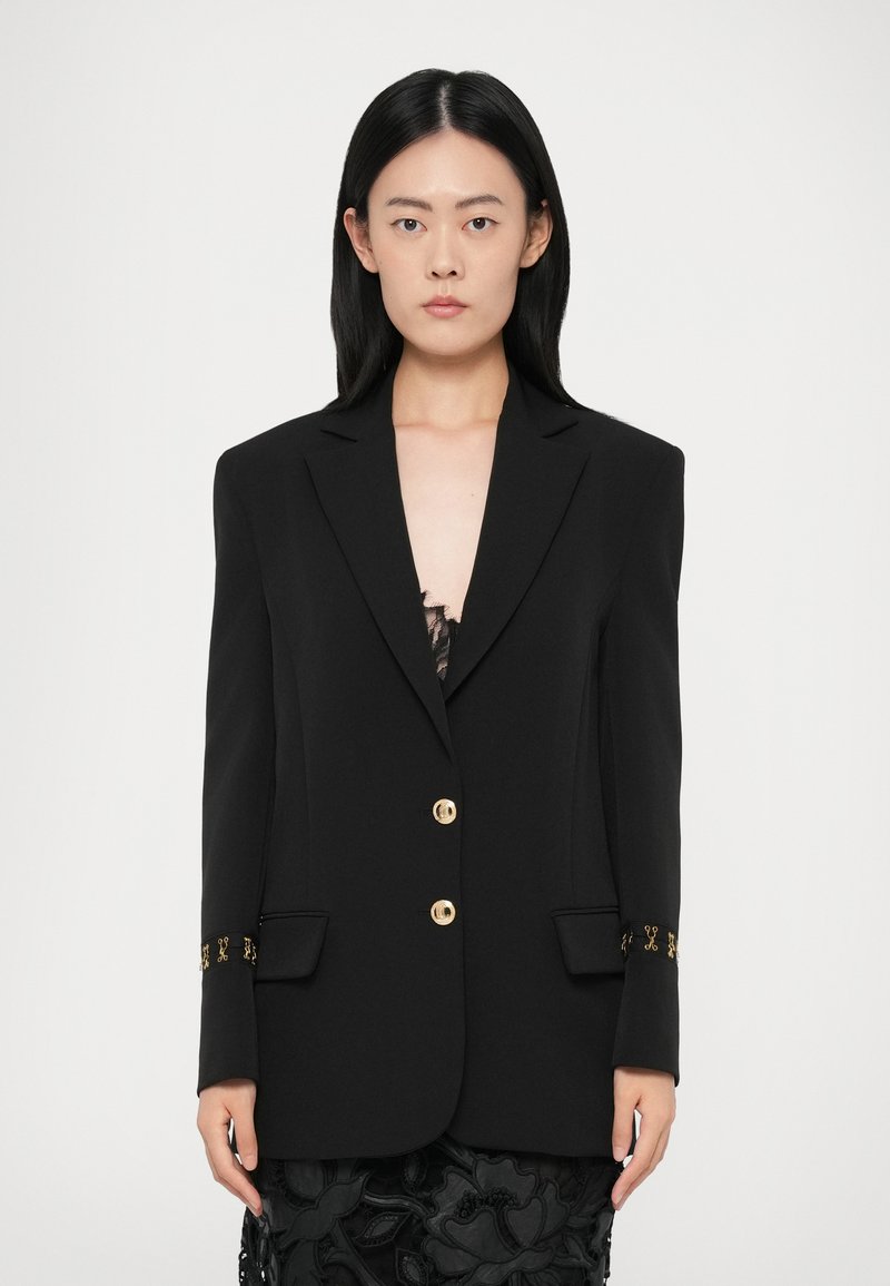Femme avec des cheveux noirs lisses portant un blazer noir avec des boutons dorés et des poignets en dentelle sur une robe en dentelle noire, devant un fond uni.