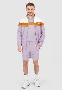 Licht paarse sportbroek met een ritsjack met witte en bruine strepen, bijpassende shorts en witte sneakers.