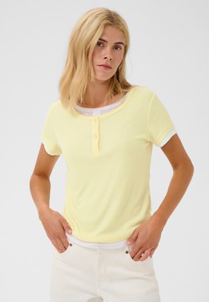 Vrouw met halflang blond haar draagt een lichtgele henley met korte mouwen en een witte jeans, staand tegen een effen lichte achtergrond.