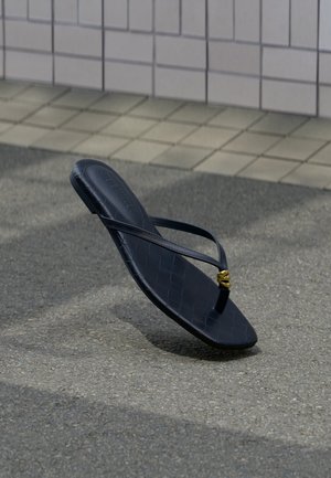 Μαύρο δερμάτινο σαγιονάρι με χρυσή διακοσμητική πόρπη, τοποθετημένο σε υφή πεζοδρομίου κοντά σε πλακιδωτό τοίχο σε εξωτερικό χώρο.
