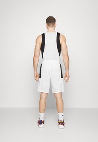 Camiseta atlética blanca sin mangas con paneles laterales negros, combinada con pantalones cortos blancos. Zapatillas multicolor y calcetines blancos hasta el tobillo visibles.