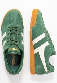 Gröna mockasneakers med vita sidostycken, snörningsdesign, gummisula och texturerad orange ytsula. Guldlogotyp på tungan.