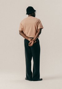 Άνδρας που φοράει μαύρο καπέλο τύπου bucket, γυαλιά, χρυσό βραχιόλι, ροζ πουκάμισο με κοντά μανίκια και μαύρο παντελόνι, στέκεται με τα χέρια δεμένα πίσω από την πλάτη.