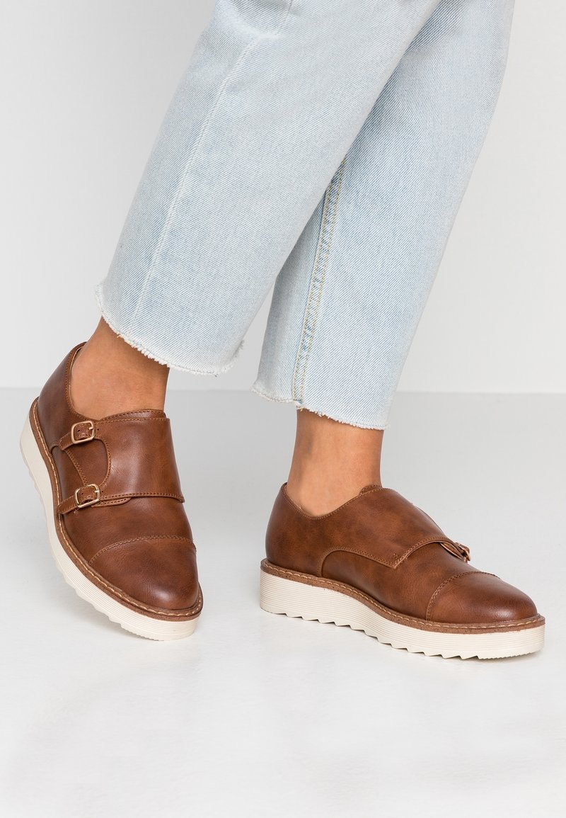 Chaussures marron en cuir à double boucle monk portées avec un jean court bleu clair sur un fond neutre.