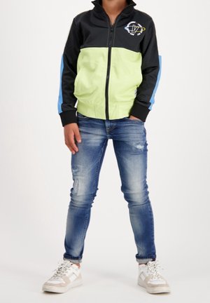 Zwart en limegroen jack met ritssluiting en blauwe accenten, gedragen over gescheurde blauwe spijkerbroek en witte sneakers. Heeft een logo op de borst.