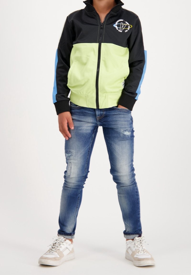 Veste zippée noir et vert citron avec accents bleus, portée sur un jean bleu déchiré et des baskets blanches. Présente un logo sur la poitrine.
