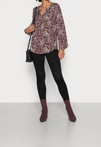 Blusa a maniche lunghe con un motivo paisley color bordeaux, abbinata a leggings neri e stivaletti bordeaux. La blusa presenta una scollatura a V.