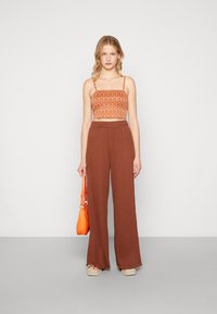 Haut orange coupé avec des motifs ondulés, associé à un pantalon large marron. Tient un sac à main orange, debout sur un fond gris clair.