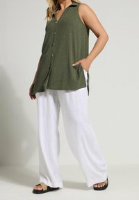 Olivově zelená bezrukávová tunika s límečkem a knoflíky, kombinovaná s bílými lněnými kalhotami s širokými nohavicemi a tmavými sandály. Viditelná jemná textura.