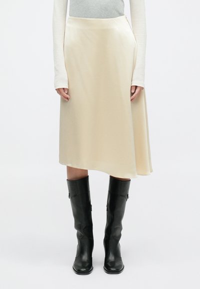 Falda A-line de satén crema con cintura alta y dobladillo asimétrico, combinada con botas de cuero negras ajustadas hasta la rodilla.