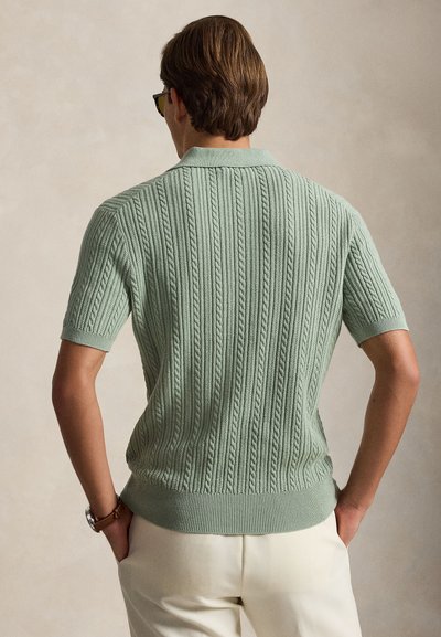 Homme portant un polo en maille torsadée vert clair à manches courtes, pantalon beige, montre et lunettes de soleil, debout, les mains dans les poches, de dos.