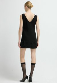 Czarna sukienka bez rękawów z dekoltem w kształcie litery V na plecach, ozdobiona misternym zdobieniem z koralików na dole, zestawiona z cienkimi czarnymi pończochami do kolan i szpiczastymi szpilkami.