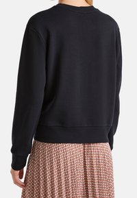 Černá mikina se širokým výstřihem a žebrovanými manžetami, s uvolněným střihem. Zadní strana se vyznačuje hladkou texturou, která kontrastuje s vzorovanou sukní.