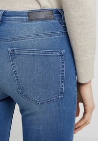 Blå denimjeans med en bakficka, som visar en läderlogg på midjan. Tyget har en lätt textur och blekning.