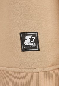 Tissu beige avec un patch rectangulaire noir affichant un logo "Starter" blanc et le texte "Black Label" cousus près d'un ourlet côtelé.