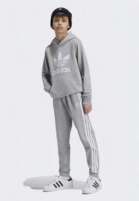 Camisola com capuz e calças de fato de treino cinza com logotipo branco da Adidas e três riscas nas laterais, combinadas com gorro preto e ténis brancos.
