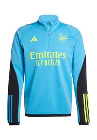Blå sportjacka med svarta ärmar, med en gul "Emirates"-logotyp och Arsenal-emblem, halvzip-design och randiga detaljer på ärmarna.