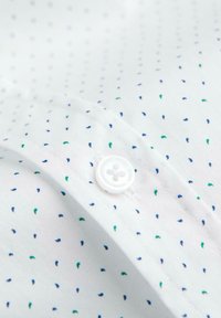 Gros plan sur un tissu blanc avec un motif de petits points bleus et verts et un bouton blanc à quatre trous cousu sur un bord plié.