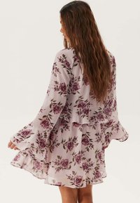 Blommig klänning i genomskinligt tyg med långa, volangärmar. Ljusrosa botten med rosor i djup burgundy och gröna detaljer.