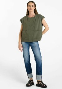 Top senza maniche verde oliva in tessuto a costine, con due tasche frontali e sei bottoni. Indossato con jeans blu scoloriti e sandali neri.