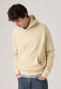 Crème-kleurige hoodie met een kangaroo zak, een capuchon met trekkoord en geribde manchetten, gelaagd over een blauw gestreept shirt.