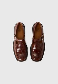 Braune Lederschuhe mit einem geflochtenen Muster, runder Spitze und einer seitlichen Schnalle. Die dicke schwarze Sohle hebt sich vom Gesamtdesign ab.