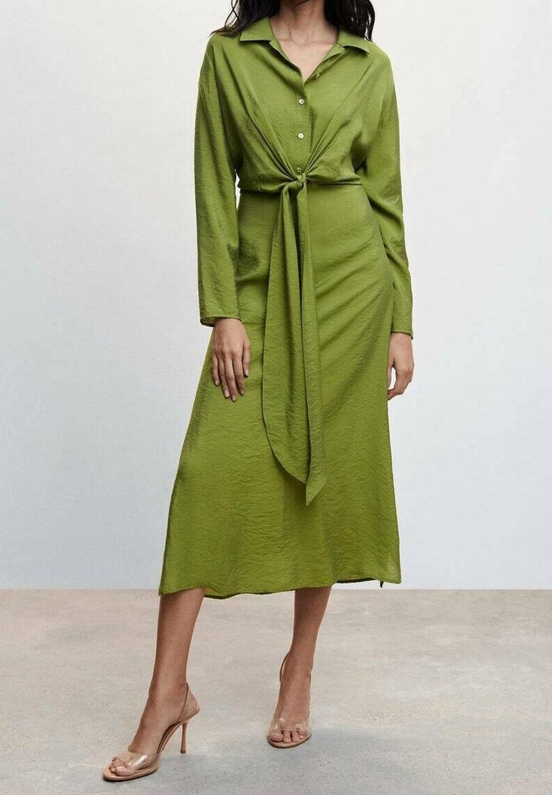Femme portant une robe-chemise verte manches longues avec un lien sur le devant, un col boutonné et des chaussures à talons beiges, debout sur un sol uni.