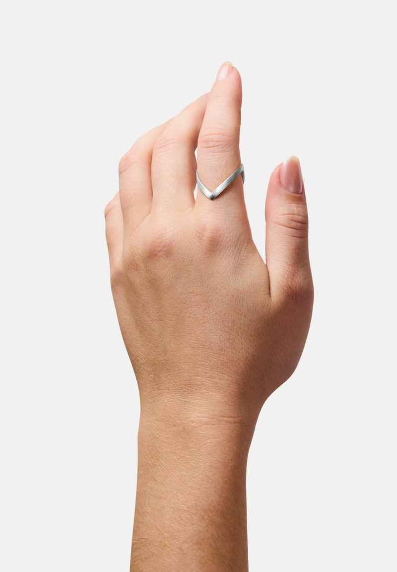Anello in metallo argento con design a chevron. La fascia è sottile e liscia, con una texture spazzolata. Mano posizionata a mostrare l'anello.