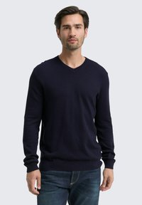 Navy V-Ausschnitt-Pullover mit gerippter Textur, langen Ärmeln und tailliertem Schnitt; getragen mit blauen Jeans. Glattes Material mit einem dezenten Glanz.