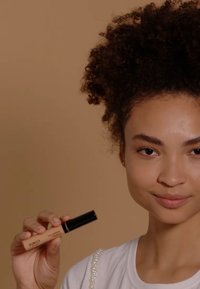 Correcteur KIKO Full Coverage dans un tube beige avec un bouchon noir, affichant le nom du produit. Le modèle a les cheveux bouclés et porte un haut blanc avec des accents en perles.