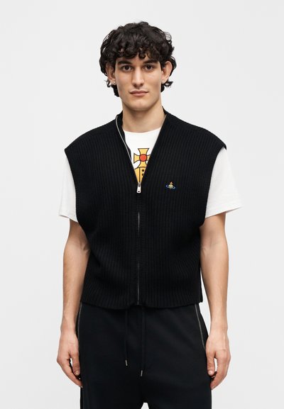 Μαύρο γιλέκο με ανάγλυφη πλέξη, φερμουάρ μπροστά, κοντά μανίκια και ένα μικρό μπλε λογότυπο στο στήθος, φορεμένο πάνω από ένα λευκό μπλουζάκι με γραφικά.